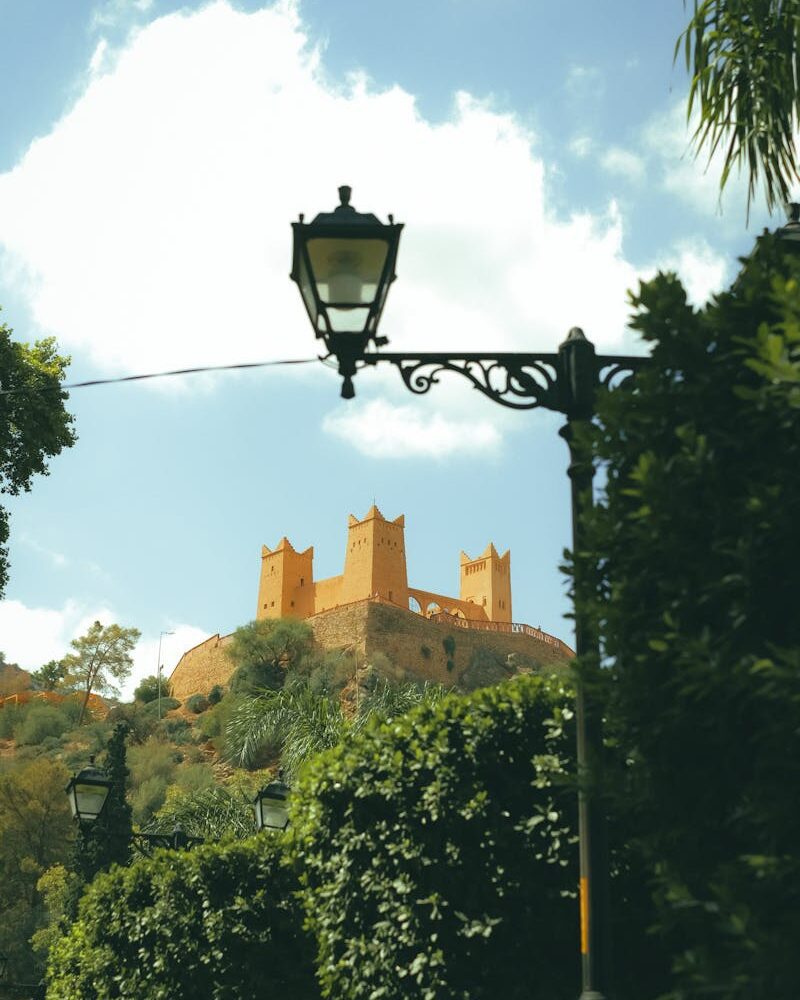 A beautiful castle on a hill with lush shrubs in Morocco, framed by a lamp post and trees.