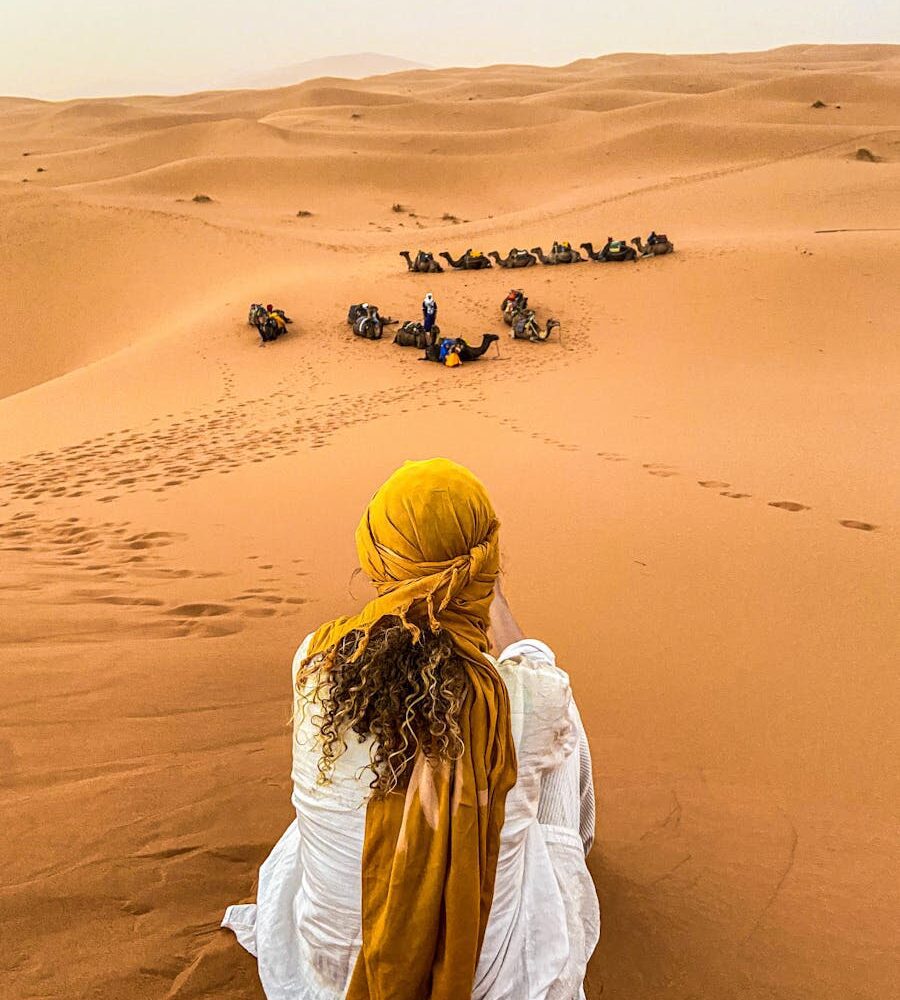 A woman gazes over a camel caravan in the Moroccan desert at sunset, serene and picturesque.